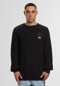 Maglione nero a maglia con texture a coste, scollatura rotonda e maniche lunghe. Presenta dettagli in suedè nero sulle spalle e una piccola etichetta con logo.