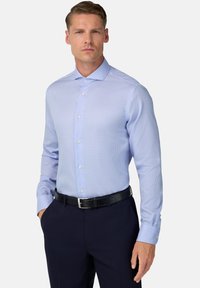 Muž v svetlom modrom kockovanom košeli zasunutej do tmavomodrých nohavíc s čiernym opaskom, stojí na pozadí s jednoduchým svetlým pozadím.