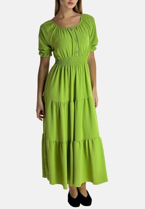 Kvinde iført limegrøn maxikjole med lag og elastisk talje, knappet rund halsudskæring, pufærmer og sorte flade sko, stående mod hvid baggrund.