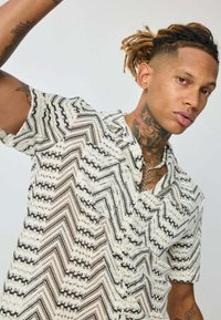 Chemise à manches courtes, à boutons, en blanc et noir avec un motif en zigzag. Présente une coupe décontractée et un tissu texturé, mettant en valeur des coutures complexes.