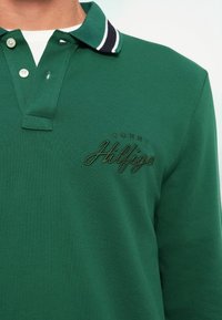 Vihreä polo-paita, jossa on teksturoitu kangas, rannetuksena tummansiniset raidat kauluksessa ja tummanvihreä brodeerattu "Tommy Hilfiger" -logo.