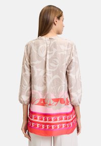 Leichte Bluse mit langen Ärmeln in Beige mit floralem Muster, versehen mit einem geometrischen Saum in Pink und Rot sowie gerafften Manschetten. Glatte Textur.