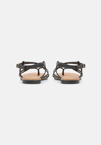 Black strappy sandals with a flat sole, featuring a tan insole, adjustable ankle strap, and gold-tone buckle detail.
