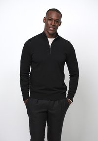 Schwarzer Reißverschluss-Pullover mit hohem Kragen und gerippten Bündchen, über einem hellen Hemd getragen. Das Modell steht lässig mit den Händen in den Taschen.