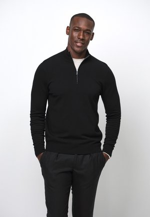 Maglione nero con zip e colletto alto, indossato sopra una camicia chiara. Il modello sta in modo informale con le mani nelle tasche.