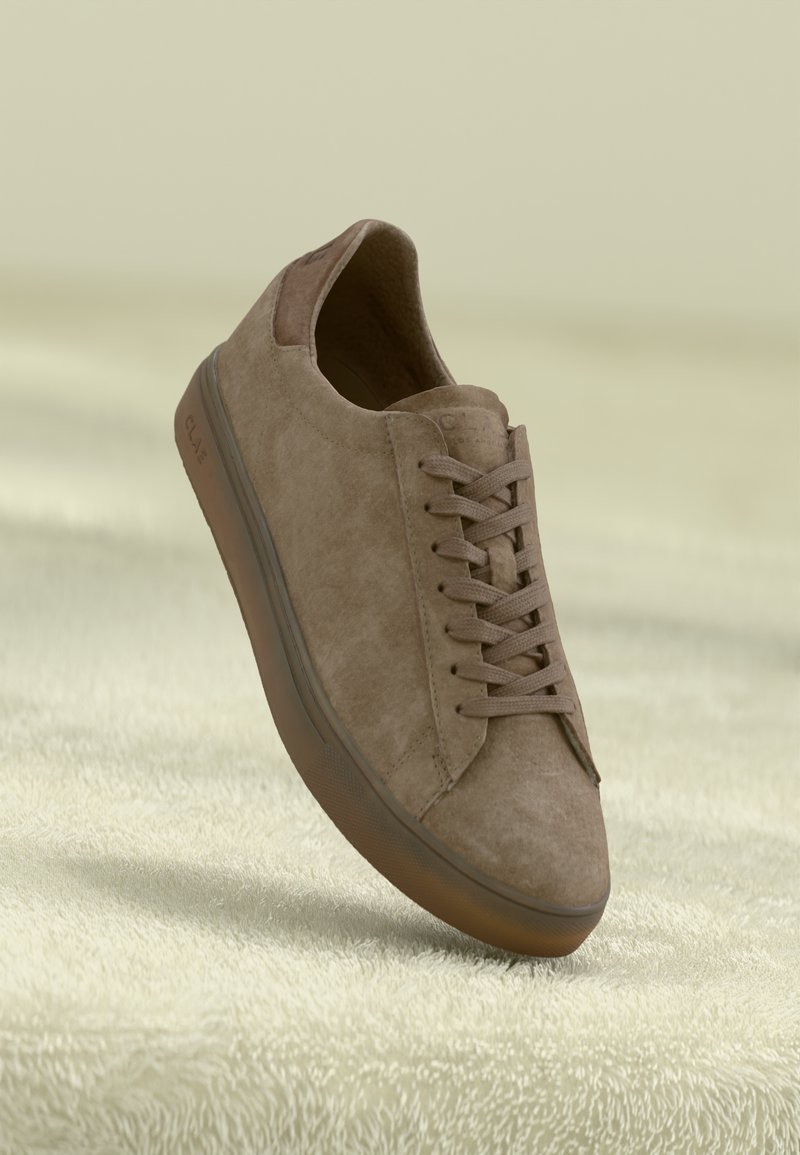Smėlio spalvos suede sportiniai bateliai su suapvalintu pirštu, toniniai raišteliai ir struktūruota guma. Yra rudas akcentas ant kulno ir reljefinė logotipas.