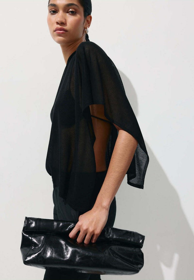 Femme portant un chemisier noir transparent aux manches longueur coude, tenant une grande pochette en cuir noir pliée devant un mur uni.