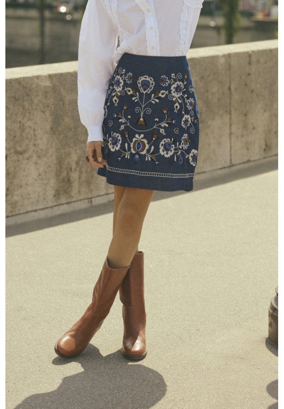 Jupe en denim avec broderies florales en bleu, blanc et vert, associée à des bottes en cuir marron jusqu'au genou. Blouse blanche avec des détails en dentelle.