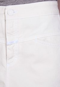 White denim pants feature a front design with a button closure, a small branded label, and contrasting white stitching.