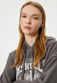 Grijze hoodie met een grote witte grafische print op de voorkant. Voorzien van een trekkoordkap en een relaxed fit, gedragen met zilveren ring oorbellen.