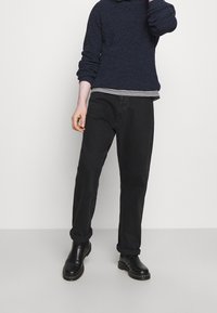 Personne portant des bottes noires, un pantalon noir retroussé, une chemise rayée et un pull bleu foncé, se tenant devant un fond clair uni.