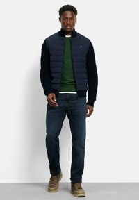 Marineblauer Puffer-Westen mit gestrickten Ärmeln über einem grünen Pullover, kombiniert mit dunklen Jeans und braunen Wanderschuhen. Verfügt über einen Reißverschluss und ein Logo.