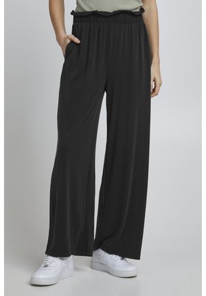 Pantalon large noir avec une taille élastique froncée, confectionné dans un matériau lisse. Dispose de poches latérales. Associé à des baskets blanches.