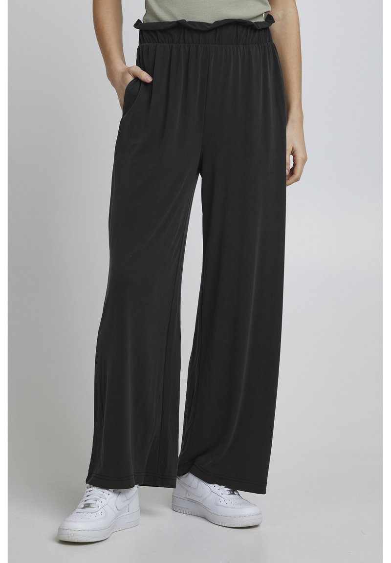 Pantalon large noir avec une taille élastique froncée, confectionné dans un matériau lisse. Dispose de poches latérales. Associé à des baskets blanches.