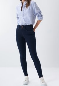 Ciemnoniebieskie, wysokie skinny jeansy z dwoma guzikami, pięcioma kieszeniami i tonowym przeszyciem. Noszone z jasnoniebieską koszulą zapinaną na guziki i białymi sneakersami.