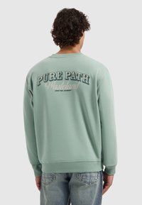 Man met krullend haar draagt een lichtgroene sweatshirt met de tekst "PURE PATH Wasteland Own The Journey" op de achterkant, gecombineerd met vervaagde blauwe spijkerbroek.