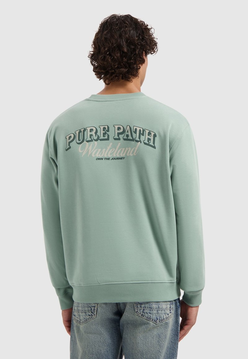 Man met krullend haar draagt een lichtgroene sweatshirt met de tekst "PURE PATH Wasteland Own The Journey" op de achterkant, gecombineerd met vervaagde blauwe spijkerbroek.