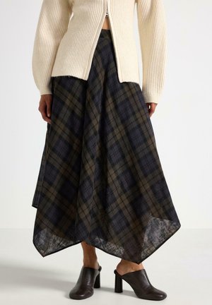 Kvinde iført en navy og brun ternet asymmetrisk midinederdel, beige ribstrikket zip-up sweater og sorte mule sko med hæl.