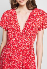 Vestido rojo con flores, con mangas cortas abullonadas, escote en V profundo y una falda fluida. Presenta pequeños patrones florales blancos y azules por toda la prenda.