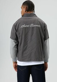 Geruit overhemd met korte mouwen in grijs en bruin met een oversized ontwerp. Voorzien van geborduurde tekst "Saint Essence." op de achterkant, gecombineerd met een grijze lange mouwlaag.