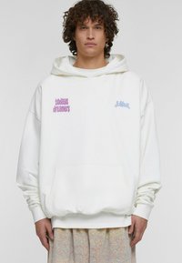 Bílá mikina s kapucí a přední kapsou, zdobená fialovým nápisem "todays dreamers" a světle modrým logem. Měkká textura, oversized design s šňůrkou na kapuci.