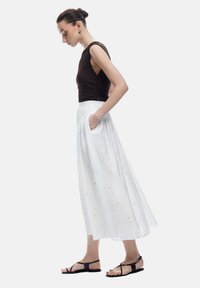 Hvid maxi nederdel med blomstermønster og lommer, parret med en brun tætsiddende top og sorte sandaler. Glat stof med et flydende design.