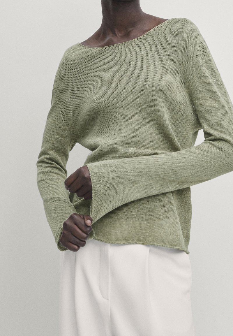 Personne portant un pull en maille vert clair ample avec des manches larges et un pantalon blanc, debout devant un fond clair uni.