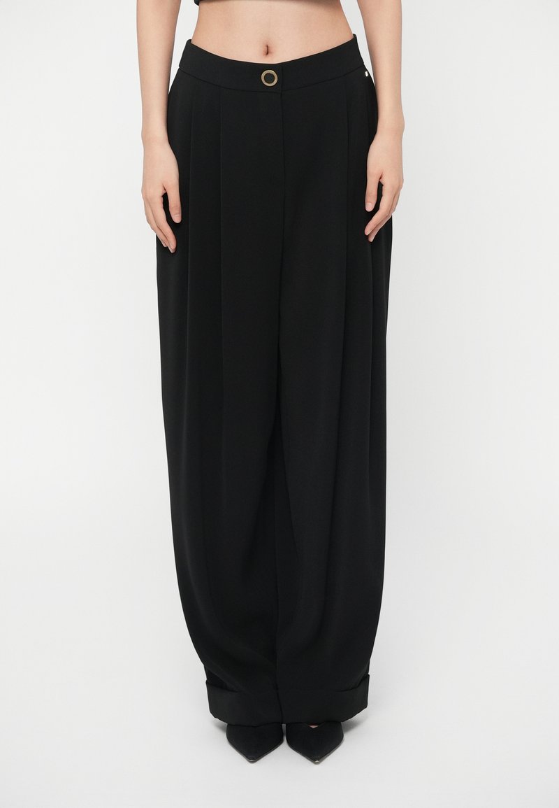 Γυναίκα που φοράει μαύρο παντελόνι με υψηλή μέση και φαρδιά πόδια, με γυρισμένα τελειώματα και χρυσό κουμπί με στρογγυλή μορφή στη μέση, καθισμένη σε λευκό φόντο.