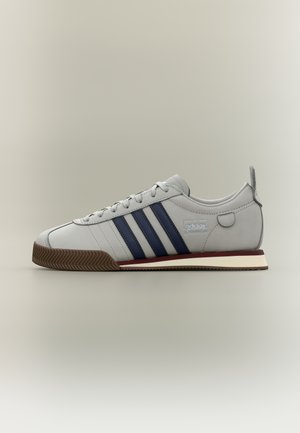 Ανοιχτό γκρι αθλητικό παπούτσι Adidas με σκουρομπλε ρίγες, καουτσούκ σόλα και μπορντό λεπτομέρεια στη μέση της σόλας, απεικονίζεται σε πλευρική προβολή σε απλό φόντο.