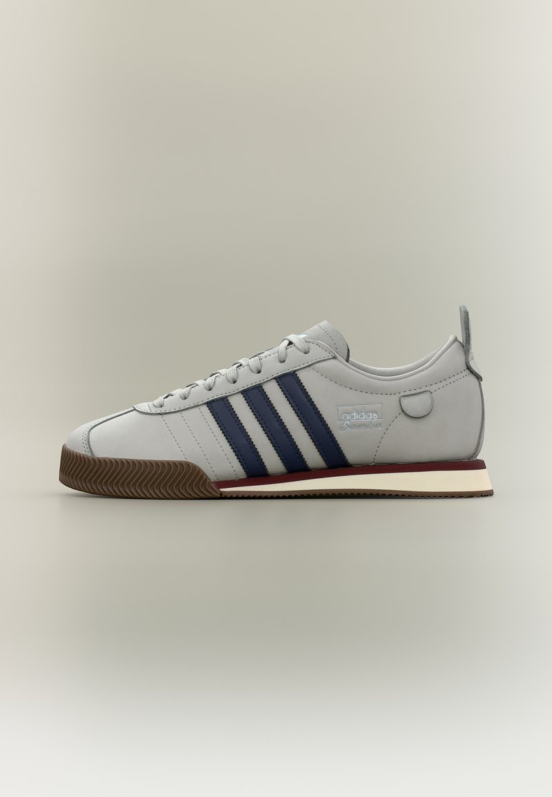 Ανοιχτό γκρι αθλητικό παπούτσι Adidas με σκουρομπλε ρίγες, καουτσούκ σόλα και μπορντό λεπτομέρεια στη μέση της σόλας, απεικονίζεται σε πλευρική προβολή σε απλό φόντο.