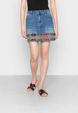 Mujer que lleva una minifalda vaquera azul con bordados geométricos de colores a lo largo del dobladillo y sandalias negras tipo slide.