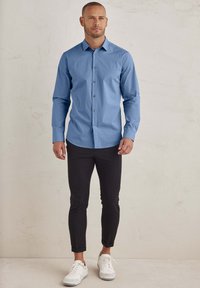 Hellblaue, slim-fit Hemd, kombiniert mit schwarzen, eng anliegenden Hosen und weißen Sneakern, vor einem neutralen Hintergrund präsentiert.