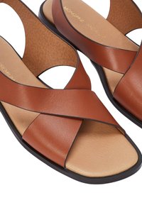 Sandales en cuir marron avec des sangles croisées, design ouvert à l'avant et semelle intérieure texturée. Semelle en caoutchouc noir, apparence minimaliste et épurée.