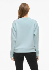 Moteris su ilgais rudais plaukais stovi, vilkėdama šviesiai mėlyną sportinį megztinį ir juodas džinsus, atsigręžusi nugara prieš paprastą baltą foną.