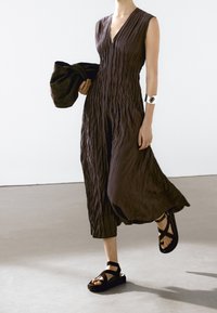 Woman walking in a sleeveless, textured dark brown midi dress, holding a matching oversized bag, wearing platform sandals and a silver cuff bracelet.