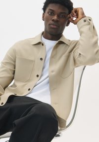 Giacca beige con colletto, due tasche sul petto, chiusura a bottoni e tessuto elegante. Indossata sopra una maglietta bianca, abbinata a pantaloni neri.