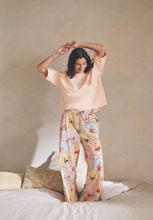 Vrouw staat op bed met opgeheven armen, draagt een licht perzikkleurige oversized top en kleurrijke bloemenprint pyjamabroek, glimlacht met gesloten ogen.