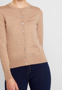 Krémový svetr vyrobený z měkké tkaniny, s kulatým výstřihem, dlouhými rukávy a stříbrným zapínáním na knoflíky vpředu. Slim fit design.