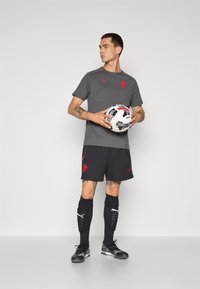 Grå atletisk t-shirt med røde detaljer, sorte shorts med røde elementer, knælange sorte sokker og sorte fodboldstøvler, mens vedkommende holder en fodbold.