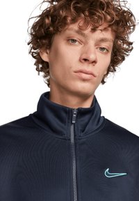 Chaqueta azul marino de cremallera con cuello alto; presenta una textura de tela suave y un logo de Nike en azul claro en el lado izquierdo del pecho.