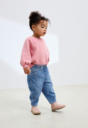 Göndör hajú kisgyermek rózsaszín pulóvert, bő kék farmert és rózsaszín bokacsizmát viselve sétál a világos padlón belül.