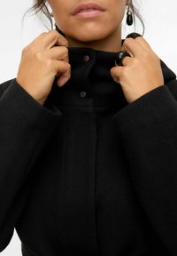 Manteau en laine noir avec col haut, fermeture à bouton-pression et épaules structuré. Présente une texture douce et des accents de design minimal.