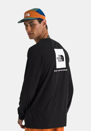 Młody mężczyzna w czarnej koszulce z długim rękawem z logo The North Face na plecach i wielokolorową czapką, patrzący przez ramię.