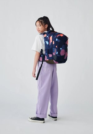 Navy-Rucksack mit leuchtenden pinken und blauen Kreismustern, gepolsterten Trägern und einer strukturierten Oberfläche. Getragen über einem weißen T-Shirt und lila Hosen.