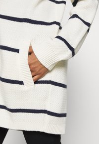Suéter blanco de punto con rayas horizontales azul marino, puños acanalados y un bolsillo delantero. Tejido texturizado de grosor medio.