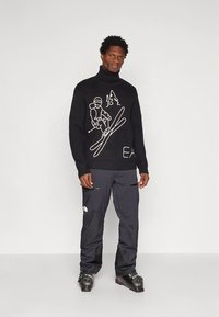 Črna pulover z visokim ovratnikom iz pletenine z belimi motivi na temo smučanja. Oblečen s temnimi izoliranimi smučarskimi hlačami in črnimi smučarskimi čevlji.