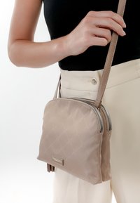 Donna che indossa un top nero e pantaloni beige, in piedi mentre tiene una borsa a tracolla beige trapuntata con doppia zip e dettaglio frangi.