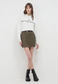 Giovane donna che indossa un hoodie bianco di Jean-Michel Basquiat, una gonna verde oliva con cintura nera e stivali militari neri con lacci, in piedi contro un muro bianco.