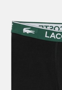 Sorte boxerbriefs med en grøn elastisk talje, der har et hvidt Lacoste-logo og et krokodillemotiv, lavet af strækbomuldsmateriale.