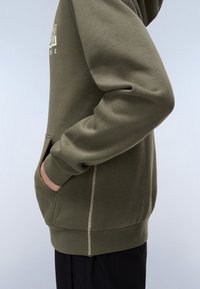 Sudadera verde oliva hecha de una suave mezcla de algodón, con un bolsillo lateral, puños acanalados y costuras blancas visibles a lo largo de las costuras.
